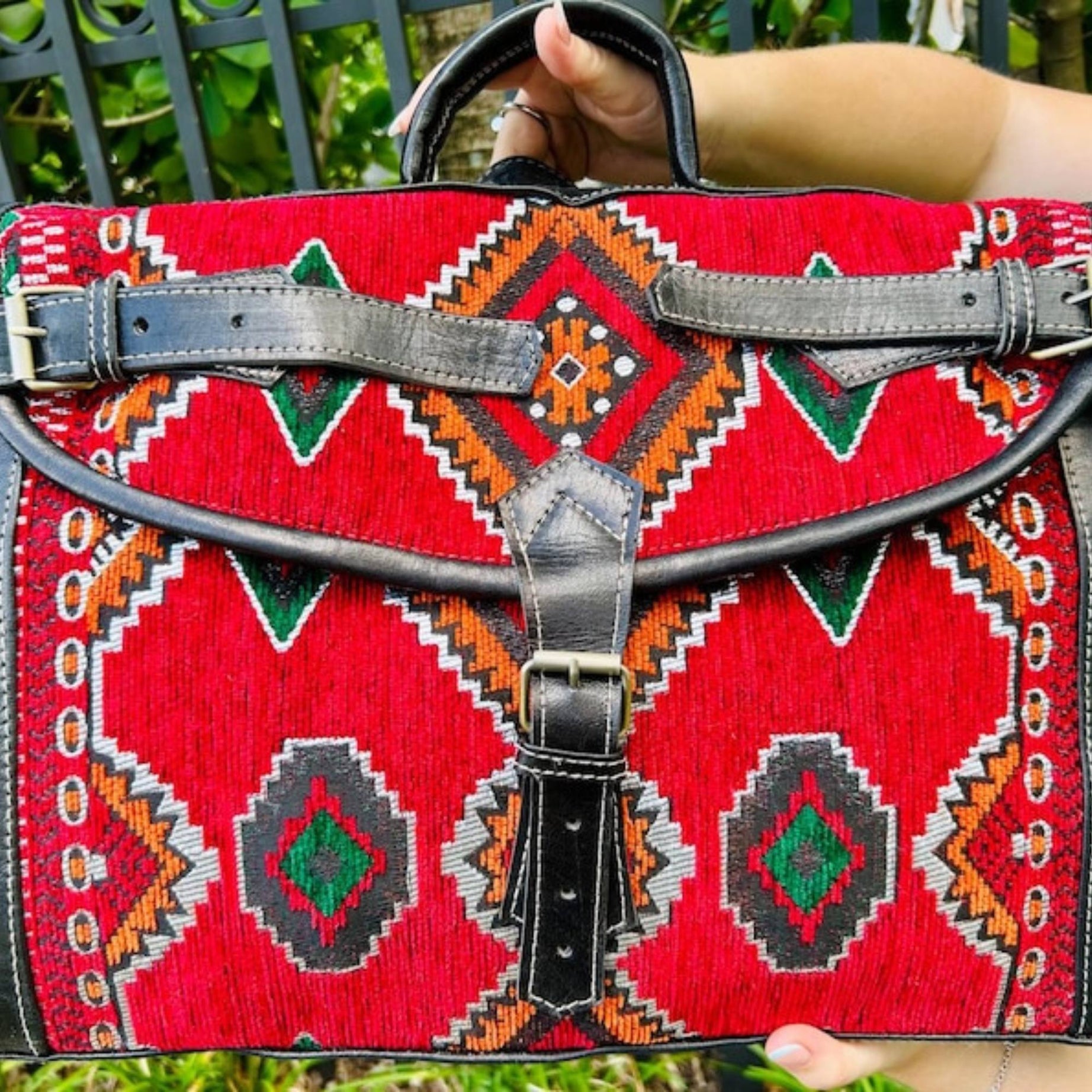 Red patterned bag with black leather straps held by a person outdoors.