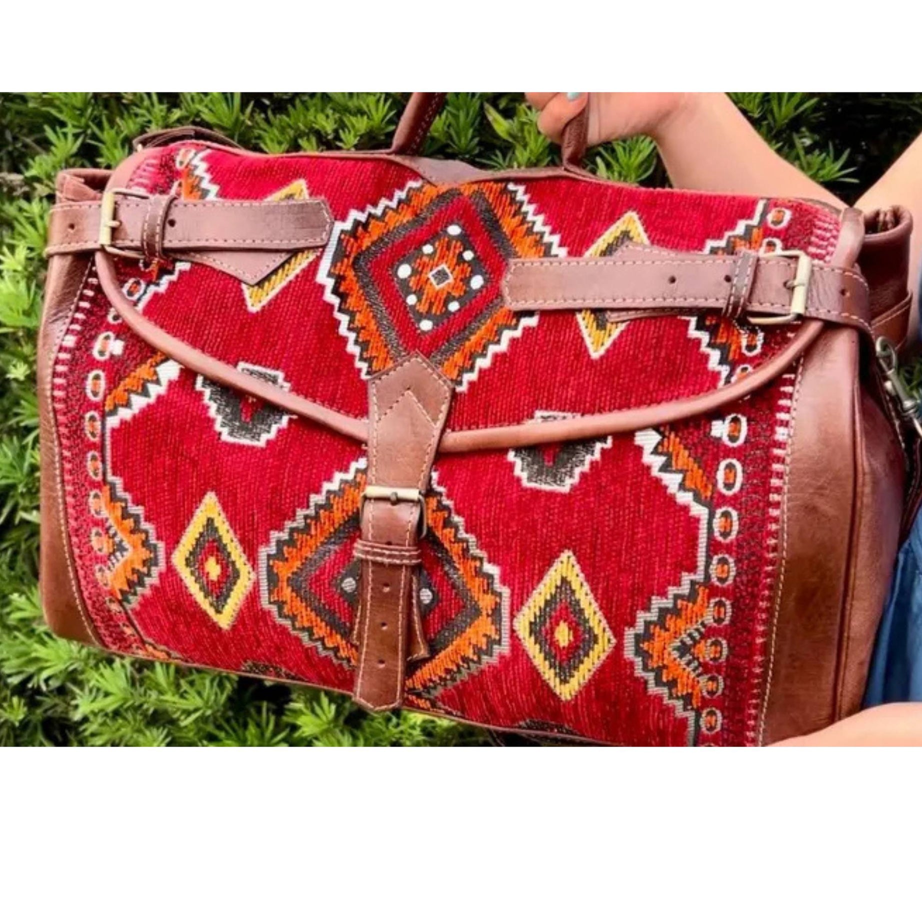 Red patterned bag with brown leather accents held by a person against a green grass background
