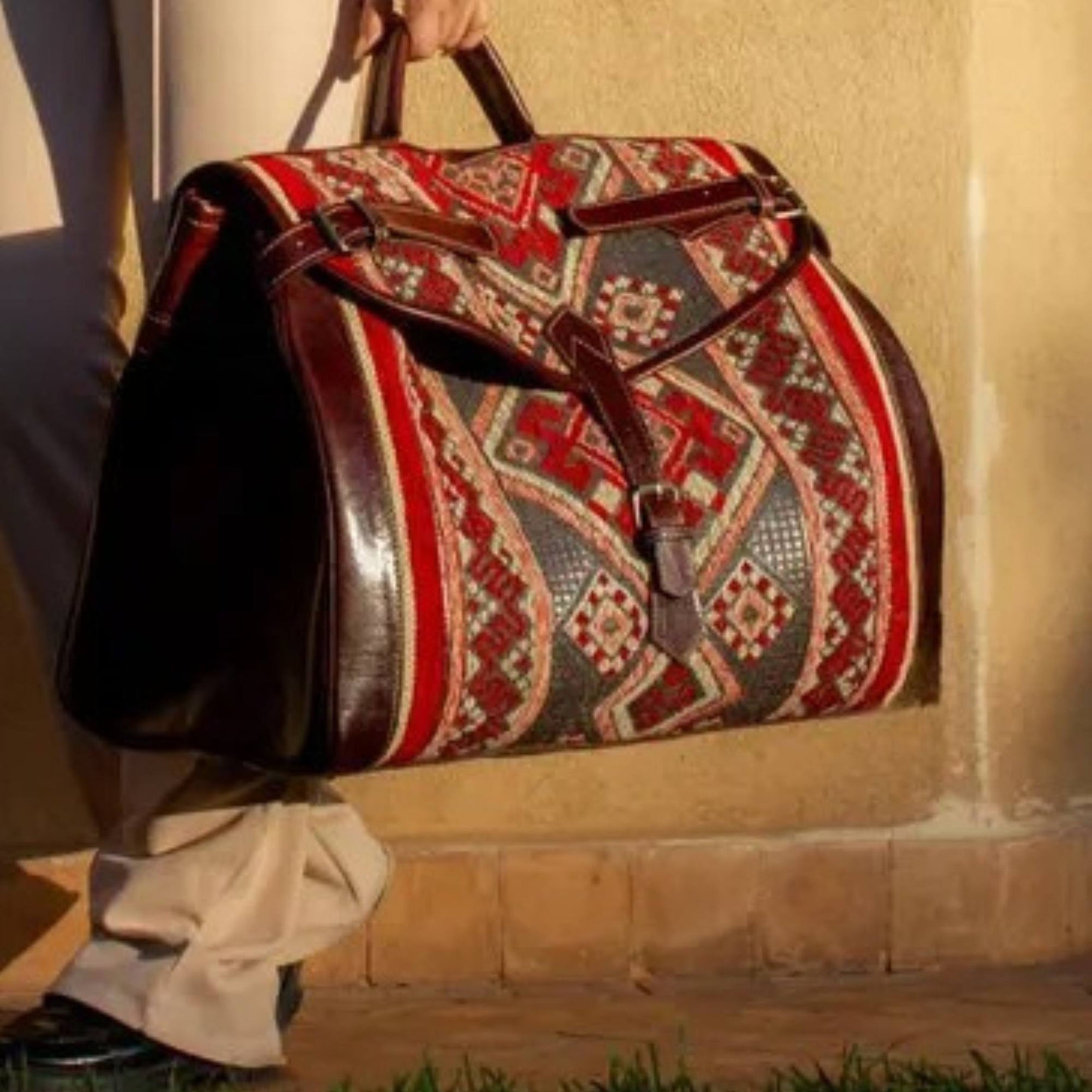 Person holding a patterned duffel bag against a stone wall.