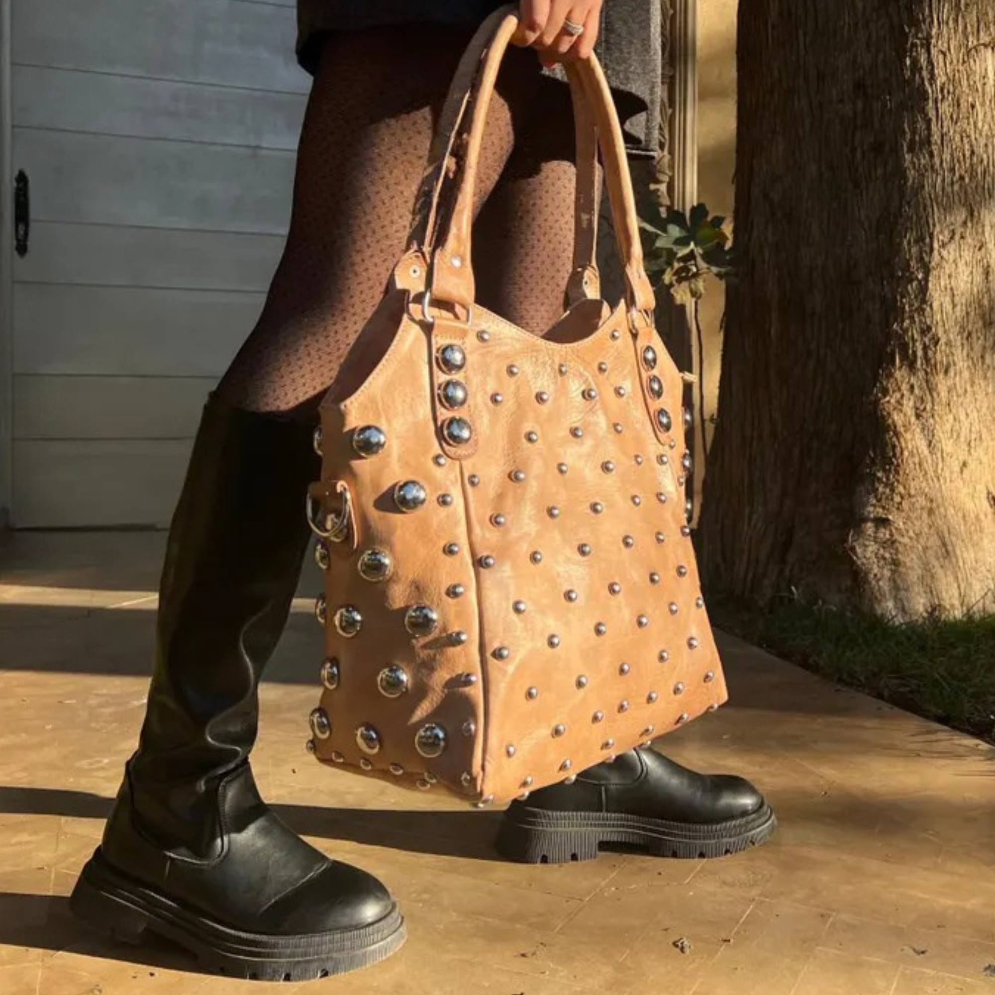 Person holding a studded tan handbag on a wooden floor.