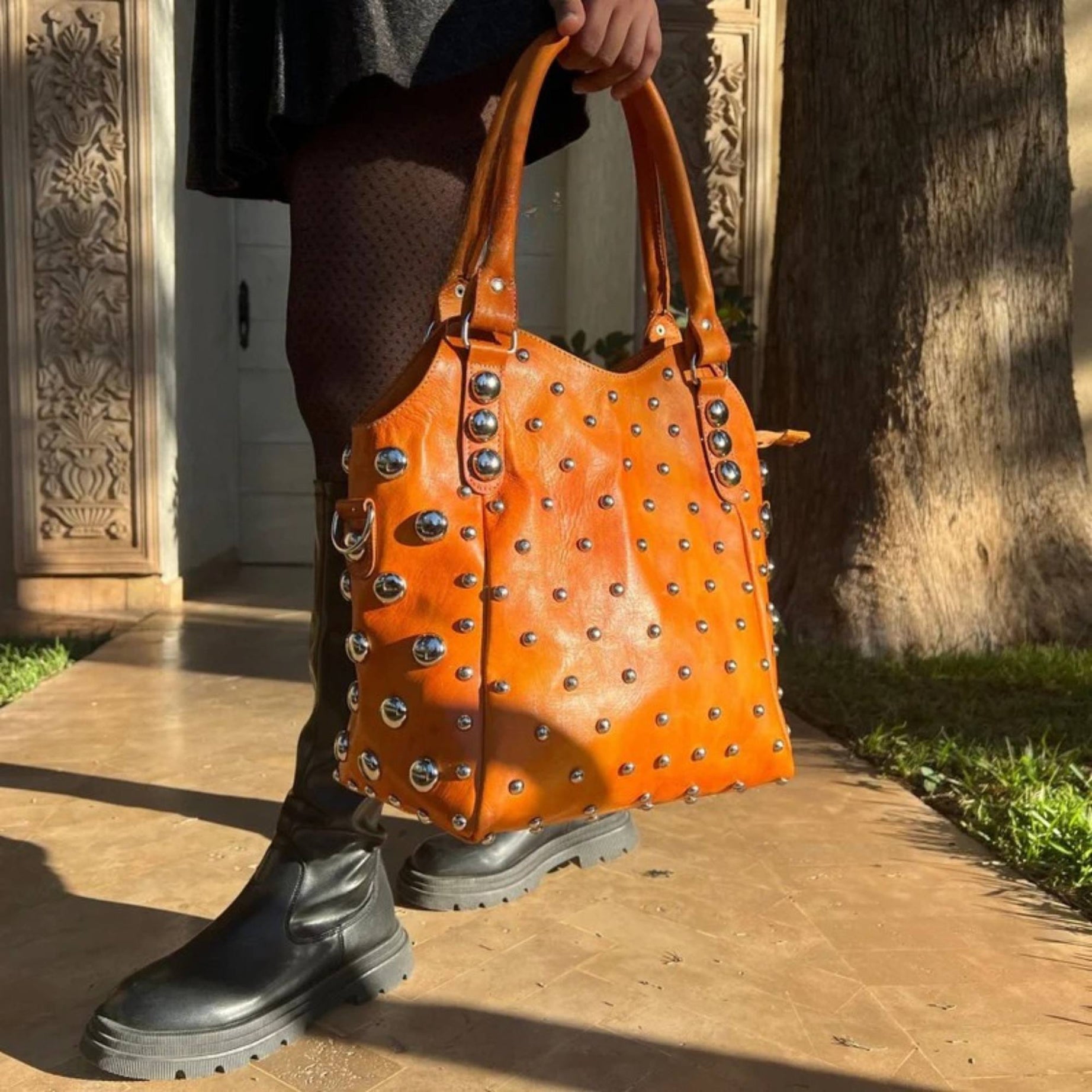 Person holding an orange studded handbag with black boots on a sunlit path.
